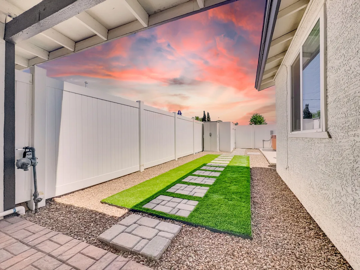 A tranquil backyard scene featuring a pathway of pavers crossing green artificial grass, bordered by a white fence and set against a vibrant, colorful sunset sky. The space is inviting for outdoor activities and family gatherings, with plenty of room for children and pets to play.