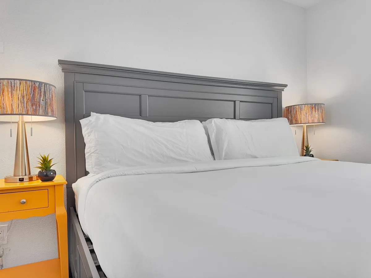 A serene master bedroom with a neatly made bed dressed in crisp white linens, a soft gray headboard, and a pop of color from the yellow bedside table. Decorative lamps on either side create a peaceful ambiance, ideal for relaxing or reading before bed.