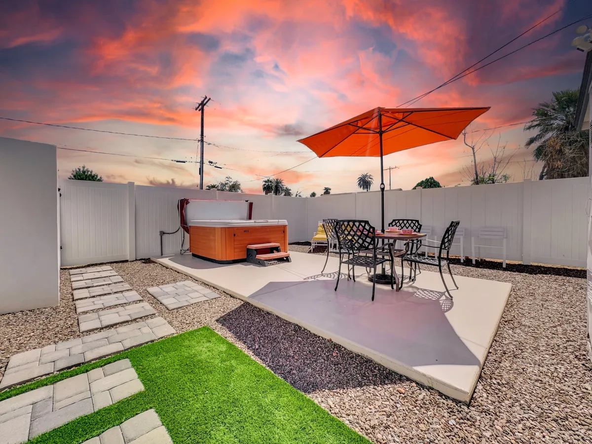 This image presents a wide view of the expansive deck, showcasing a bubbling hot tub as the main focal point. The deck is surrounded by lush greenery, offering a sense of seclusion and tranquility. An outdoor dining set with an orange umbrella sits nearby, perfect for meals or lounging. A green lawn area extends from the deck, hinting at space for pets and social gatherings.