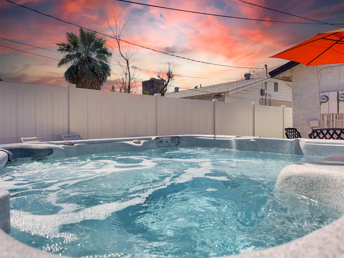A tranquil scene focuses on the hot tub, shaded by a vibrant orange umbrella and framed by a palm tree and a soft, cloud-streaked sky. The image emphasizes the hot tub as the centerpiece, inviting guests to imagine soaking in privacy while surrounded by natural beauty.