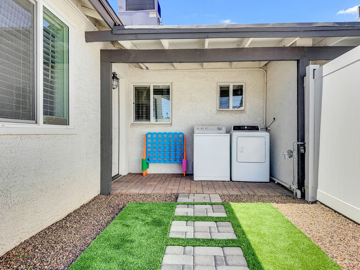 A casual exterior space with colorful outdoor seating, a pathway of stones leading through the yard, and laundry machines in view. The area feels lively and functional, with an emphasis on relaxation and family-friendly gatherings under sunny skies.