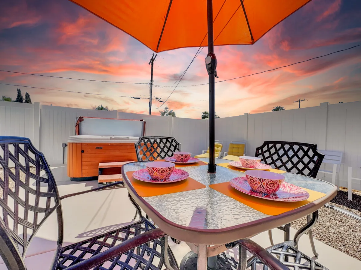 This lively image spotlights an outdoor dining table set with colorful plates and cups beneath an orange umbrella, with the hot tub visible in the background. The setting is cheerful, inviting guests and their pets to enjoy meals or celebrate special moments outside.