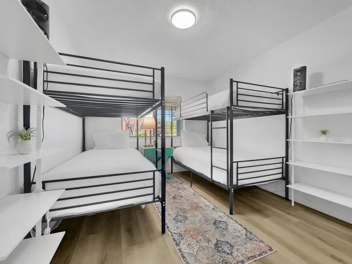 A cheerful second bedroom furnished with sturdy bunk beds, a large rug, and wall shelves for added convenience. A window lets in natural light, creating a communal and flexible sleeping space perfect for families or groups of friends.