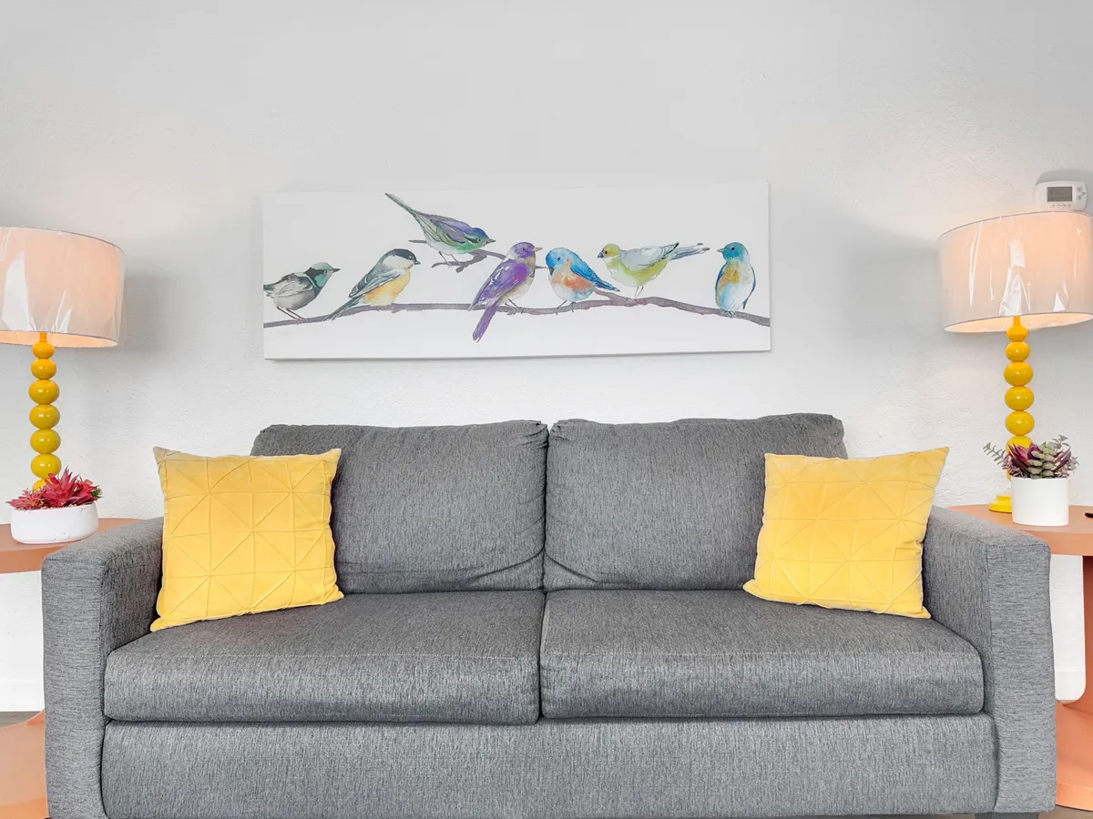 This interior scene features a cozy living room with a gray couch accented by yellow pillows, bird-themed artwork on the wall, and a pair of table lamps. The inviting space suggests a relaxed, homey atmosphere ideal for unwinding after a day out with your pet.