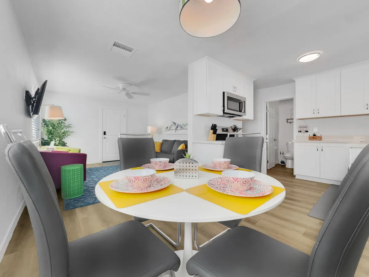 This image spotlights a round dining table set with colorful dishes and surrounded by brightly colored accent chairs, adjacent to a modern kitchen. The space is lively, cheerful, and ready for shared meals or casual gatherings.