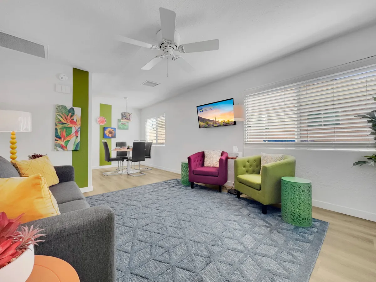 This image presents a lively living room featuring colorful accent chairs, a modern coffee table, tasteful artwork on the walls, and a plush rug. The TV on the wall and soft lighting create a space that is perfect for both lively gatherings and quiet relaxation.