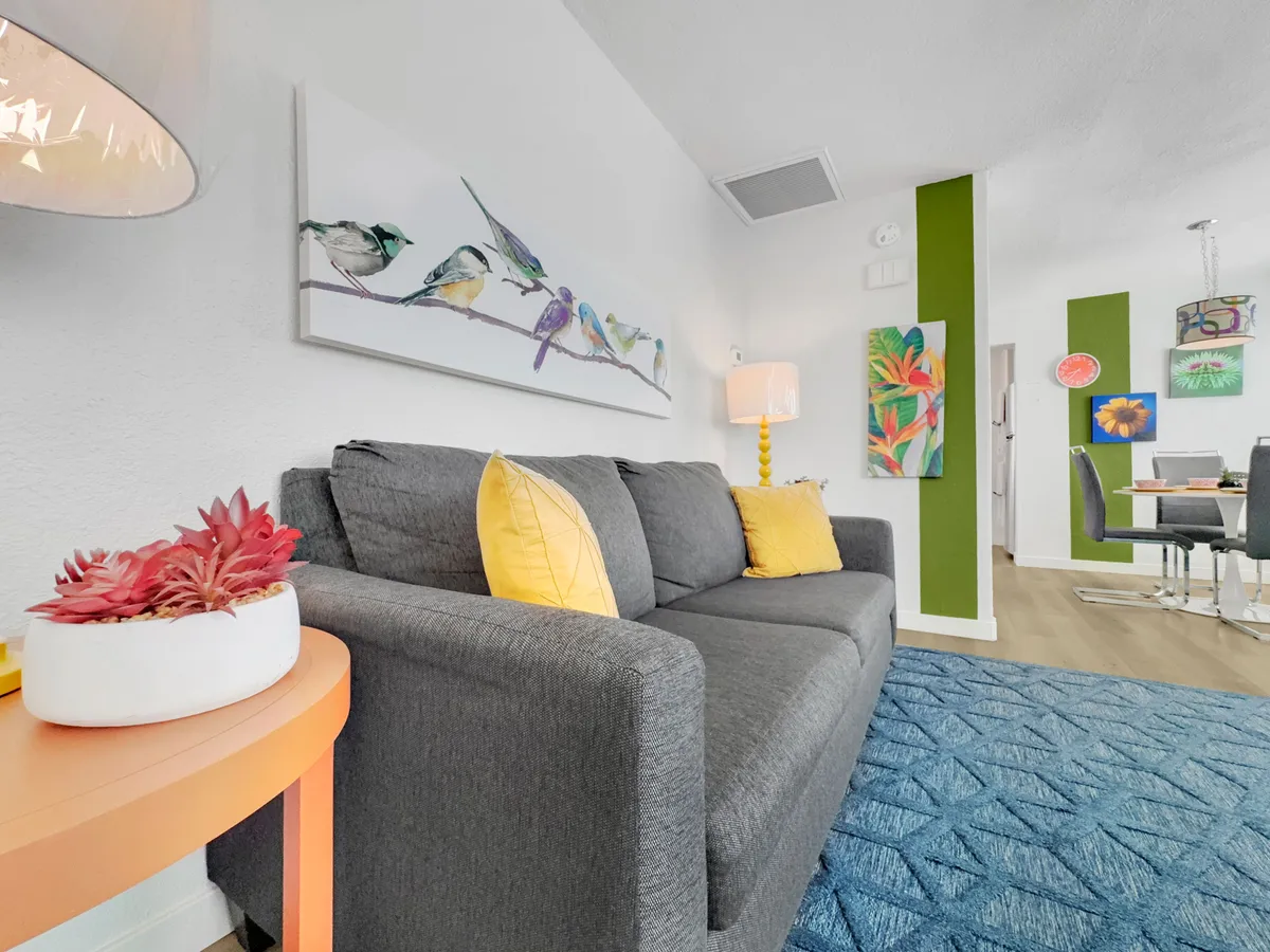A bright, open-plan view connects a gray sofa and colorful artwork with a visible dining area in the background. The green accent wall and decorative lighting create a cheerful, inviting setting that blends living and dining spaces seamlessly.